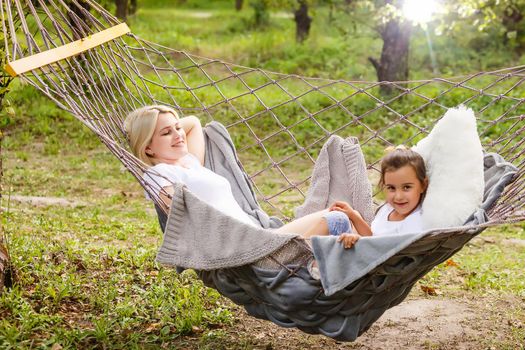 Beautiful young mother and daughter laying down and relaxing together on a hammock during a sunny summer day on holiday home garden. Family relaxing outdoors, healthy and wellness lifestyle.