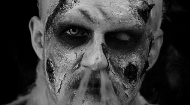 Sinister dead man close-up. Zombie guy with make-up with wounds scars and white contact lenses blows smoke from mouth, smiles terribly, trying to scare in dark studio room. Halloween, filming, staging