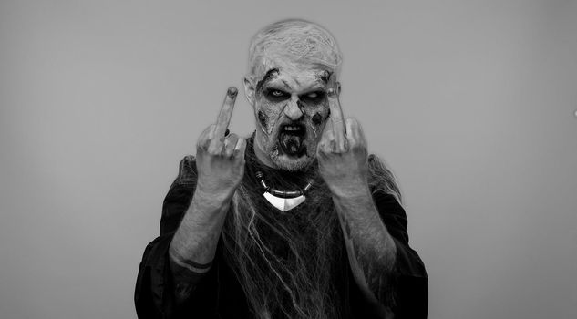Aggressive crazy hooligan Halloween zombie man with makeup with wounds scars showing around his middle fingers, demonstrating protest with impolite rude gesture of disrespect, rejecting communication