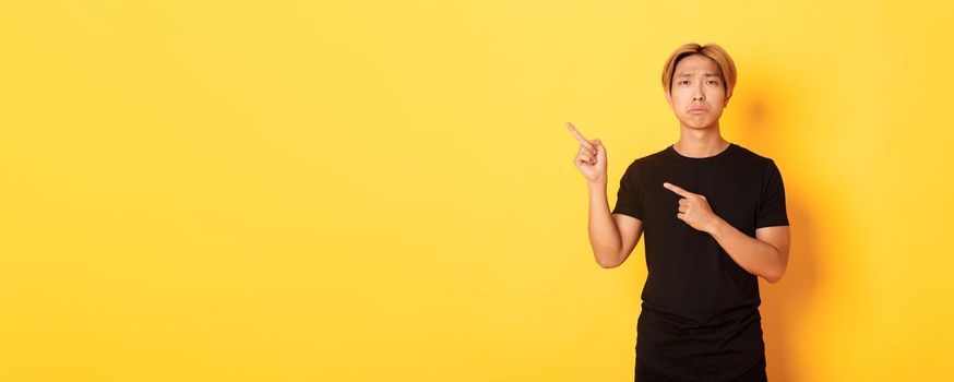 Portrait of misearble and gloomy asian guy in black t-shirt, pointing fingers upper left corner disappointed, yellow background.