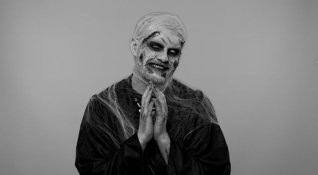 Sneaky cunning zombie man with makeup with wounds scars with tricky face gesticulating and scheming evil plan, thinking over devious villain idea, cunning cheats, jokes and pranks. Sinister dead guy