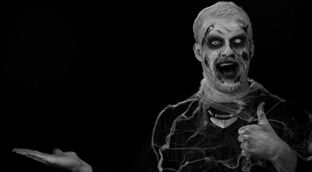 Zombie man with makeup with fake wounds scars showing thumbs up and pointing empty place, advertising area for commercial text logo, copy-space for goods promotion. Sinister Halloween ugly dead guy