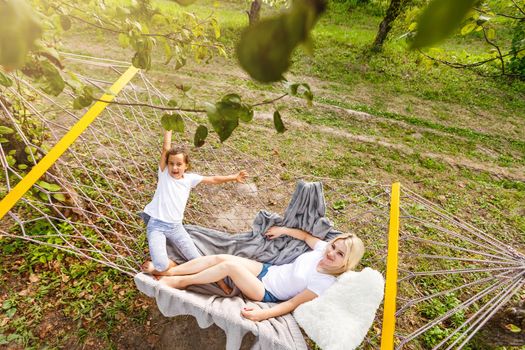 Happy mother and daughter relaxing together in a hammock at garden in sunny summer day. Family playing in hammock.