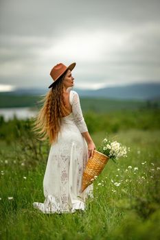 A middle-aged woman in a white dress and brown hat stands on a green field and holds a basket in her hands with a large bouquet of daisies. In the background there are mountains and a lake