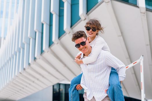 Carefree young urban couple in sunglasses doing piggyback at city street outdoor with modern commercial building on background. Man carrying young woman on his back, positive emotion have fun walking