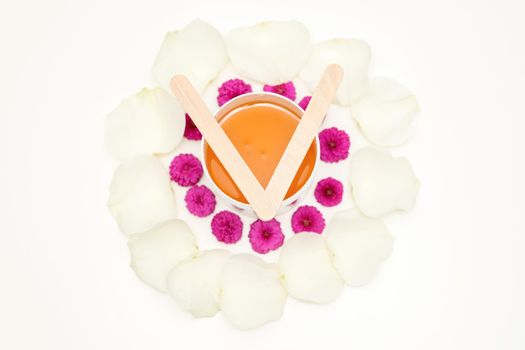 Jar of wax with wooden sticks, pink flowers, and white petals on white background. Concept of depilation. Flat lay top view