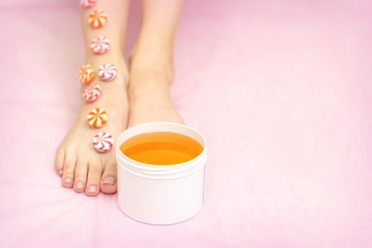 Round candies lying in a row on female feet with white jar with sugar paste on pink background with copy space, depilation concept