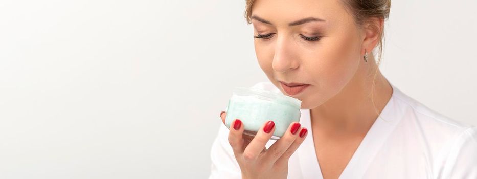 Portrait of young beautician sniffing moisturizing cream holding white jar on white background, copy space