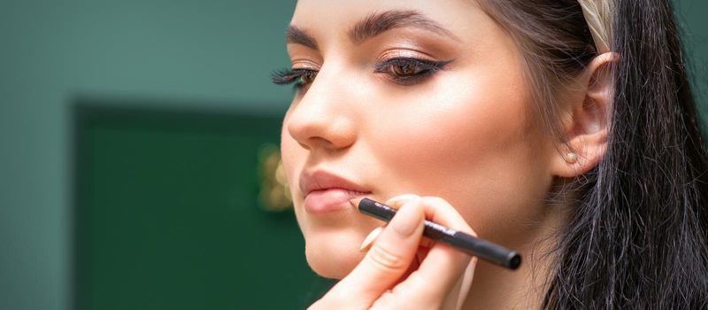 Makeup professional artist or cosmetologist is painting contour lips of a young woman with a pencil close up