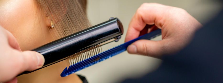 A hairdresser is straightening the hair of the young brunette woman in a beauty salon