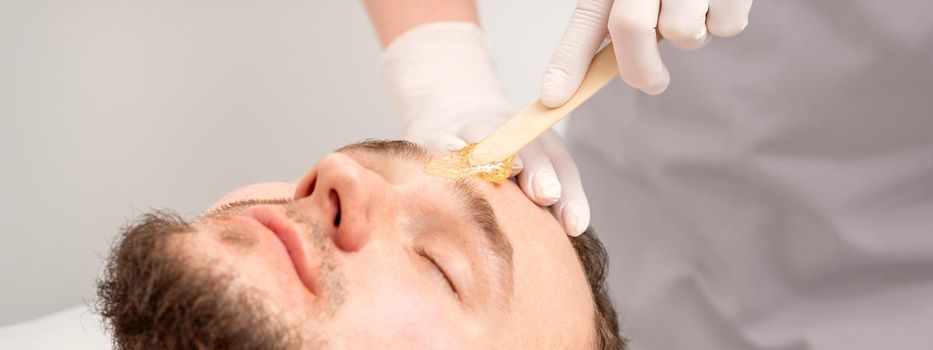 Beautician applying wax paste between eyebrows during the procedure of waxing in the beauty salon