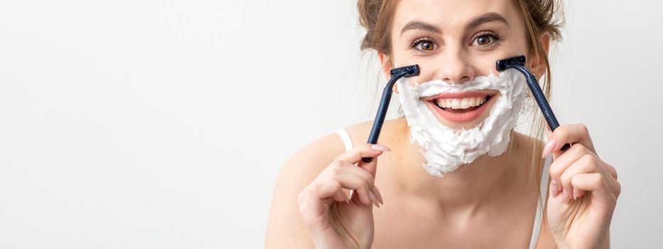 Beautiful young caucasian woman shaving her face by razor on white background. Pretty smiling woman with shaving foam and razor on her face