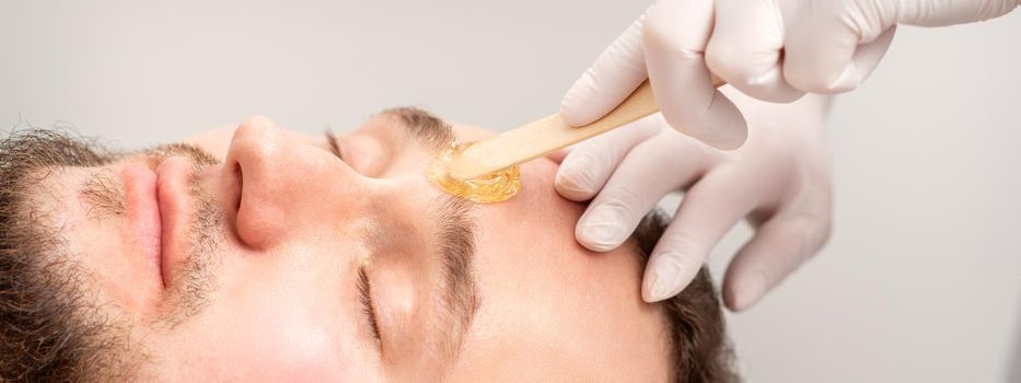 Beautician applying wax paste between eyebrows during the procedure of waxing in the beauty salon