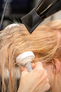 Hairdresser hand drying blond hair with a hairdryer and round brush in a beauty salon