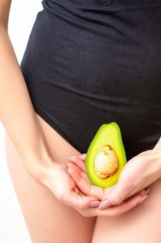 Young woman holding one half of a fresh avocado fruit close to her belly against a white background, healthy nutrition and pregnancy concept