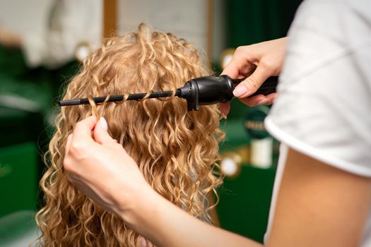 Hands of hairstylist curl wavy hair of young woman using a curling iron for hair curls in the beauty salon rear view