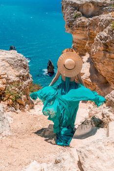 A girl with loose hair in a long mint dress descends the stairs between the yellow rocks overlooking the sea. A rock can be seen in the sea. Sunny path on the sea from the rising sun.