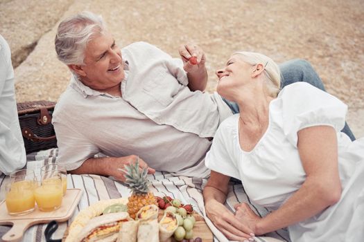 Food, beach and picnic senior couple dating or on romantic honeymoon date with fruit, snack and drink. Happy, love and romance woman and man or elderly people eating and feed together for anniversary.