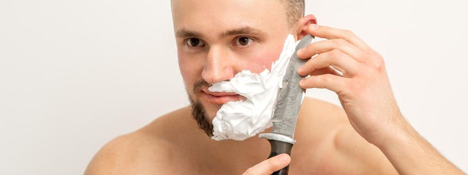 Young caucasian man shaving beard with a big knife on white background