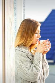 Woman relaxing on the balcony with coffee. A beautiful young woman in a sweater holds a cup of coffee standing near the window in the morning