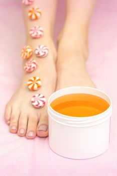 Round candies lying in a row on female feet with white jar with sugar paste on pink background with copy space, depilation concept