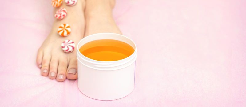 Round candies lying in a row on female feet with white jar with sugar paste on pink background with copy space, depilation concept