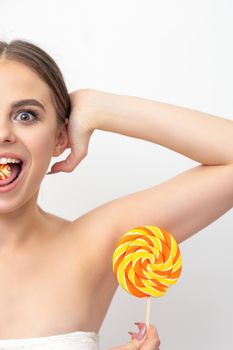 Happy expression young nude woman with candy in mouth holds lollipop near armpit on white background, epilation, depilation, hair removal concept