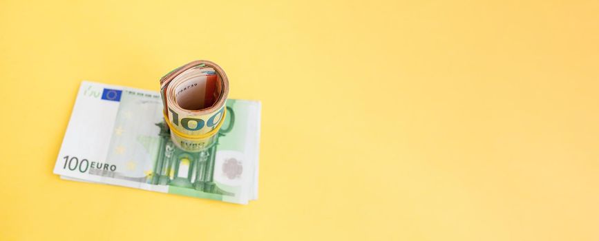 Money roll of euro banknotes with a elastic band on a 100 euro paper bill on a yellow background. cash currency, payment, earnings and savings, the concept of money and finance. Empty space for text