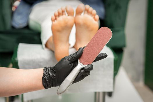 Special grater for scrubbing heels in hands of nail pedicure technician on feet background in beauty salon closeup