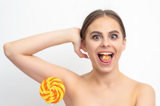 Happy expression young nude woman with candy in mouth holds lollipop near armpit on white background, epilation, depilation, hair removal concept
