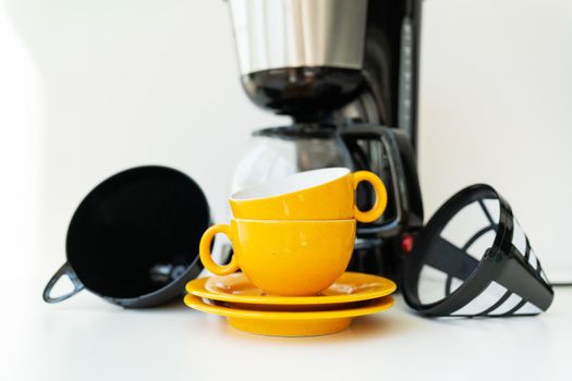 An automatic drip coffee maker stands with a yellow cup on a white table. Black modern drip coffee pot. Electric kitchen small household appliances. Appliances