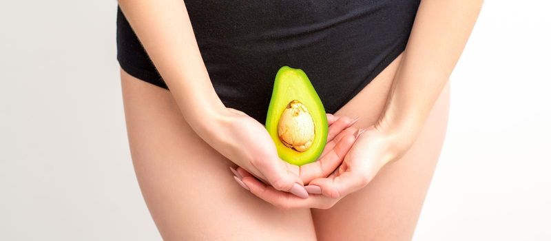 Young woman holding one half of a fresh avocado fruit close to her belly against a white background, healthy nutrition and pregnancy concept