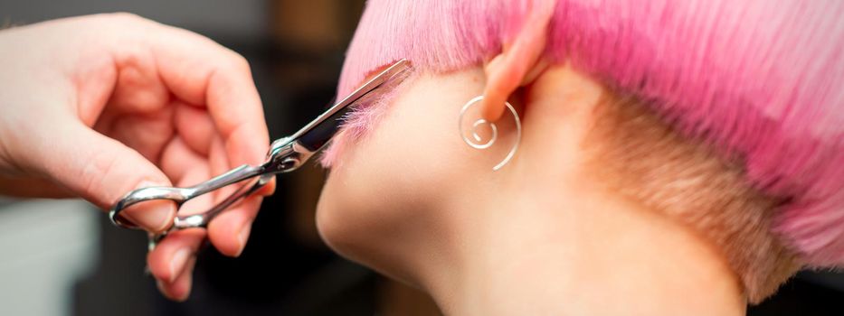 Cutting female hair. Hand of a hairdresser cutting short pink hair of young white woman at the hair salon