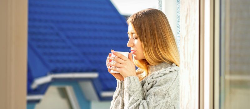 Woman relaxing on the balcony with coffee. A beautiful young woman in a sweater holds a cup of coffee standing near the window in the morning