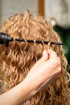 Hands of hairstylist curl wavy hair of young woman using a curling iron for hair curls in the beauty salon rear view