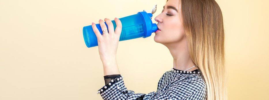 Thirsty woman with blue plastic bottle against a light background. A beautiful young caucasian businesswoman in a blouse drinking water on yellow