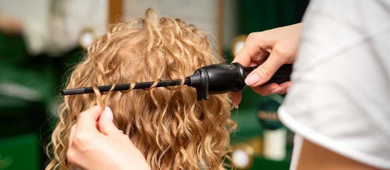 Hands of hairstylist curl wavy hair of young woman using a curling iron for hair curls in the beauty salon rear view