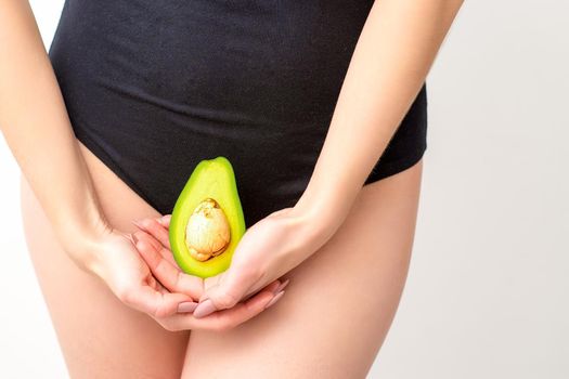 Young woman holding one half of a fresh avocado fruit close to her belly against a white background, healthy nutrition and pregnancy concept