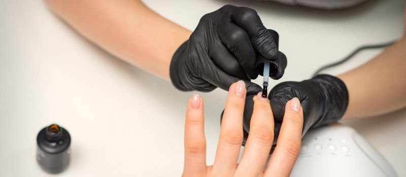 Painting nails of a woman. Hands of Manicurist in black gloves applying transparent nail polish on female Nails in a beauty salon