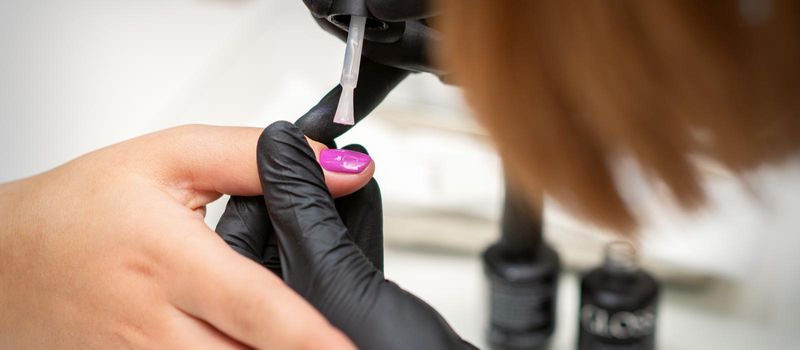 Painting nails of a woman. Hands of Manicurist in black gloves applying pink nail polish on female Nails in a beauty salon