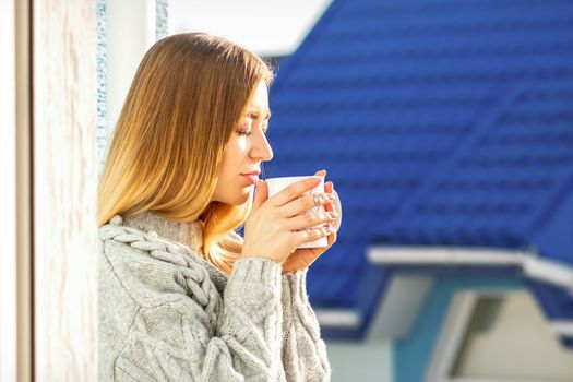 Woman relaxing on the balcony with coffee. A beautiful young woman in a sweater holds a cup of coffee standing near the window in the morning