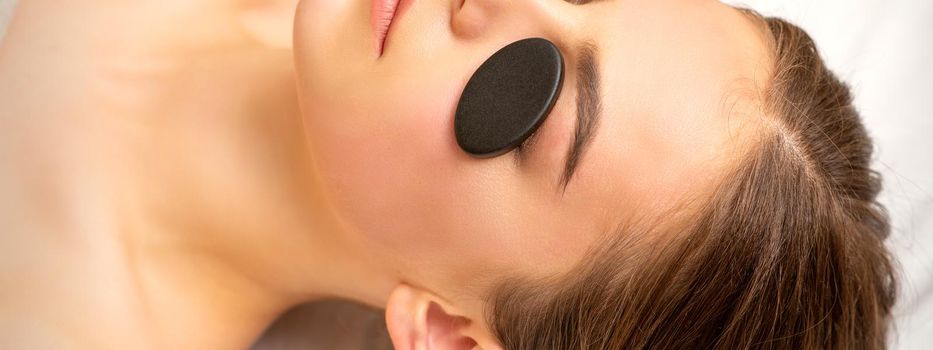 Black massage stones lying on the eyes of the young caucasian woman. Facial massage in a spa