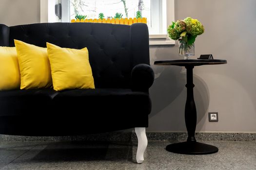 Interior of the beauty room. Black sofa with small yellow pillows and coffee table on a gray wall background with a window