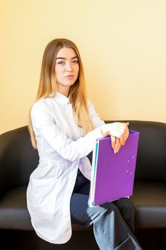 The young healthcare professional, dentist, doctor, nurse, the assistant in a white coat with folders sitting on a black sofa and looking at the camera