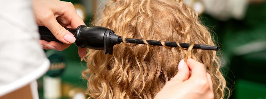 Hands of hairstylist curl wavy hair of young woman using a curling iron for hair curls in the beauty salon rear view