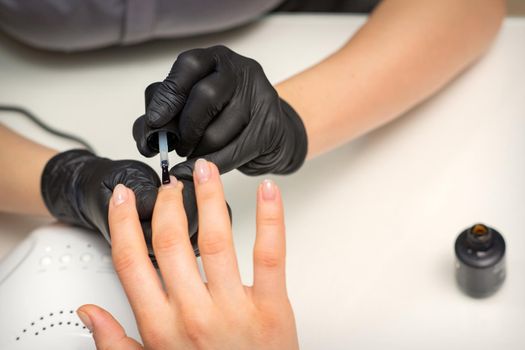 Painting nails of a woman. Hands of Manicurist in black gloves applying transparent nail polish on female Nails in a beauty salon