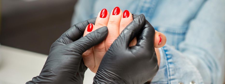 Examination of manicured fingernails. Hands of manicure master in black gloves examining female red nails in manicure salon