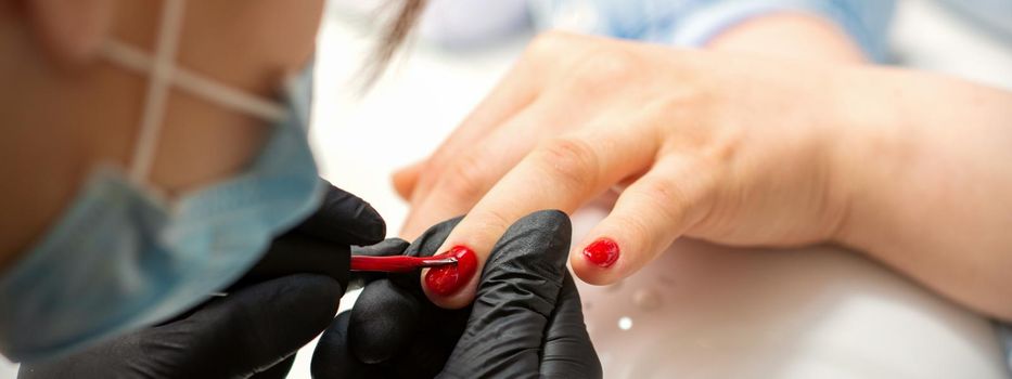Painting nails of a woman. Hands of Manicurist in black gloves applying red nail polish on female Nails in a beauty salon