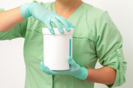 Hands in protective gloves of beautician open a white body cream jar on white background