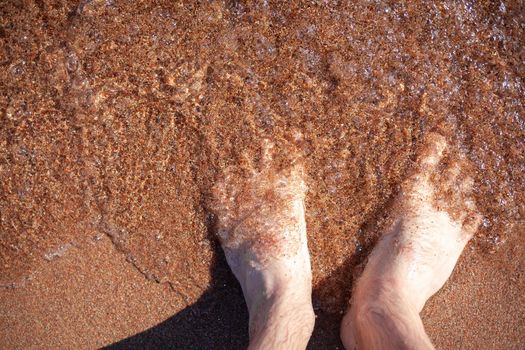 Top view of men's legs in the sea. A man is standing on the sand. An ocean wave washes the shore. Summer holidays.